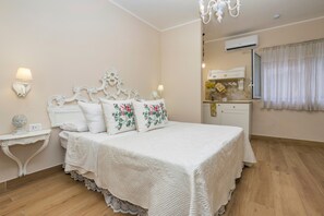 A bedroom with a bed, bedside lamps, a nightstand, a chandelier, an air conditioning unit, and a window with curtains.