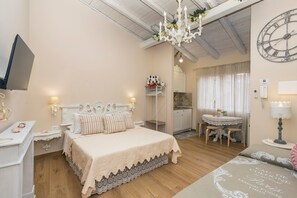 A bedroom with a bed, bedside tables, a TV, a chandelier, a clock, a small table with chairs, and a bathroom visible in the background.