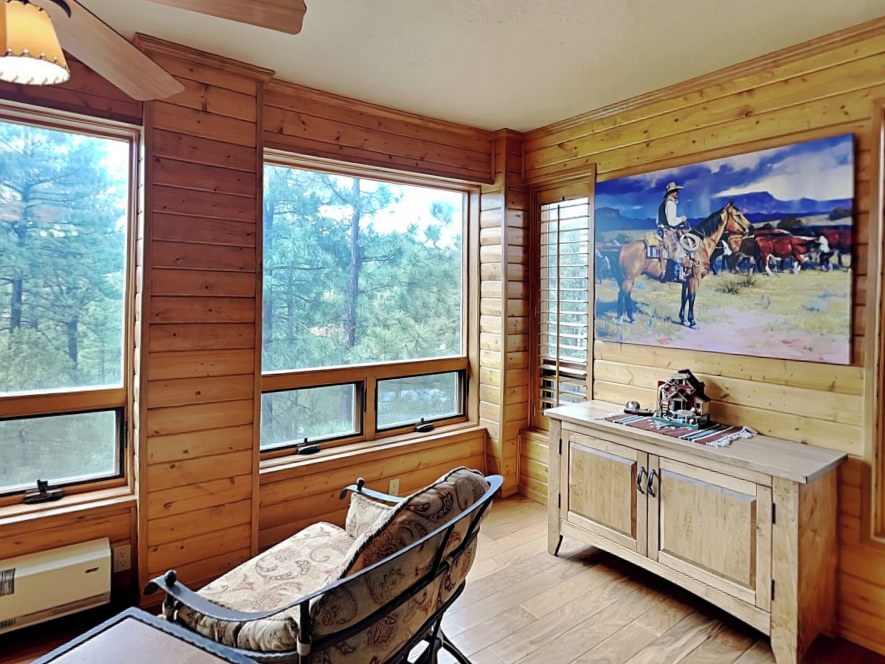 A wooden cabin interior with large windows, a dining table, a cushioned chair, a cabinet, and a painting of a cowboy scene.