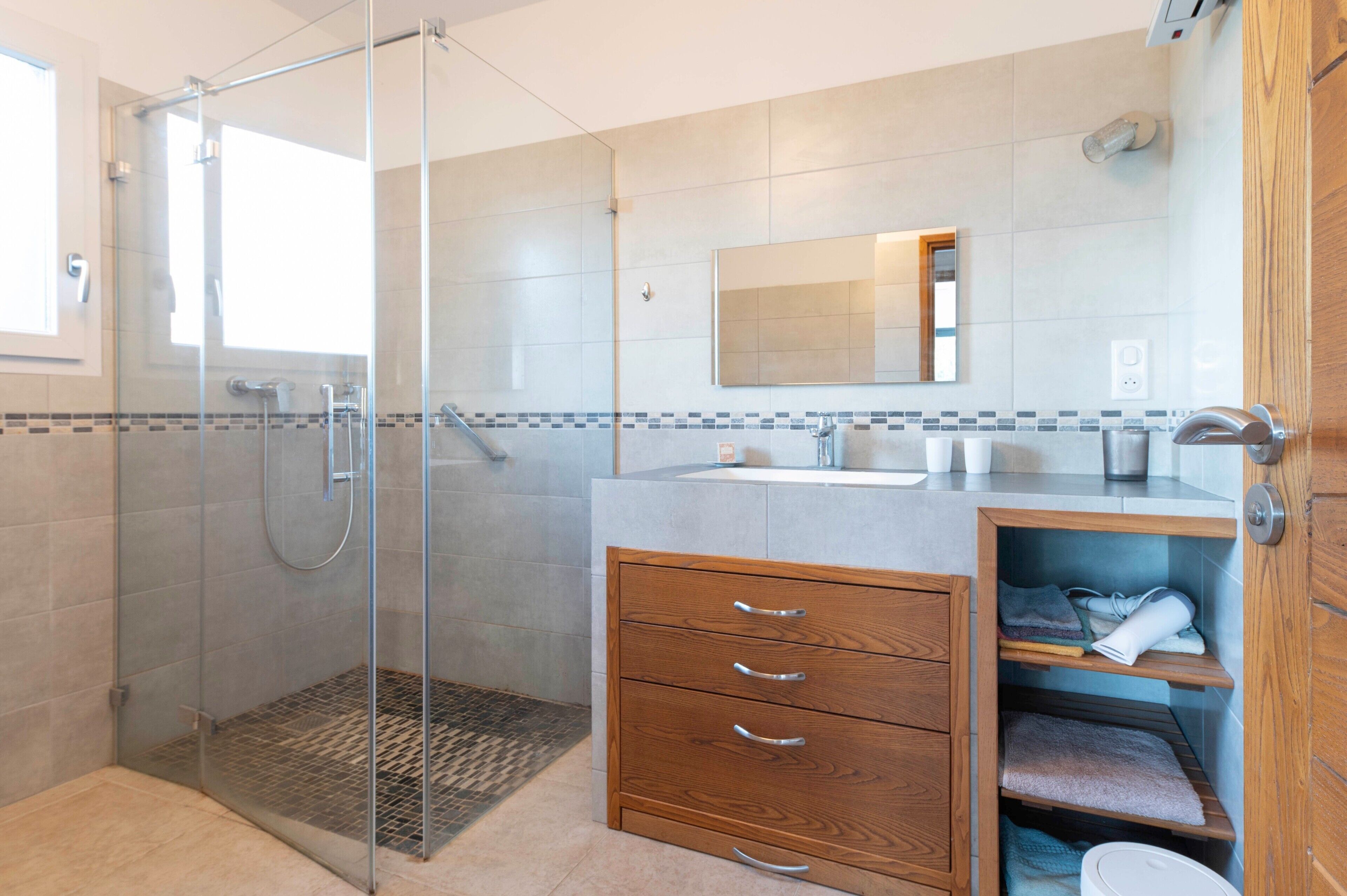 Een moderne badkamer met een douchecabine van glas, een houten wastafelmeubel met lades, een spiegel en een betegelde spatwand.