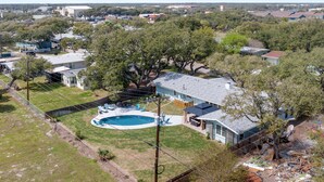 An aerial view of a residential area with a large house featuring a swimming pool, surrounded by trees and other houses.