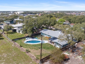 An aerial view of a residential area with a large house featuring a swimming pool, surrounded by trees and other houses.