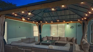 An outdoor patio with wicker furniture and a fire table, under a covered area with string lights.
