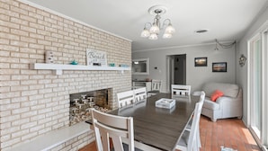 A dining area with a brick wall, white chairs, a wooden table, and a fireplace.
