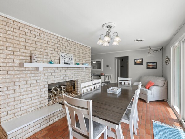 A dining area with a brick wall, white chairs, a wooden table, and a fireplace. - The Sea Glass House by Miss Kitty’s (Rockport)
