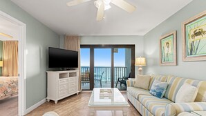 A living room with a TV, a sofa, a coffee table, and a balcony with a view of the sea. - 702 | Cozy Fall Beachfront Retreat—1BR, Gulf Views, Pool & VIP Beach Service (Panama City Beach)