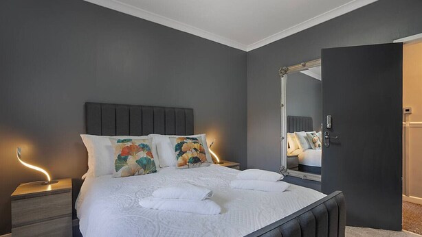 A neatly made bed with white linens and decorative pillows, a large mirror, and bedside lamps in a modern bedroom with dark walls. - Hilton Road Apartment (Aberdeen)