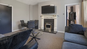 A modern living room with a fireplace, a flat-screen TV mounted above it, a dining area with chairs, and a sofa. - Hilton Road Apartment (Aberdeen)