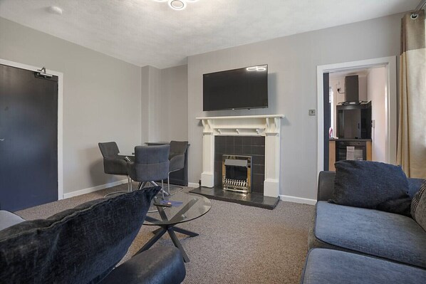 A modern living room with a fireplace, a flat-screen TV mounted above it, a dining area with chairs, and a sofa. - Hilton Road Apartment (Aberdeen)
