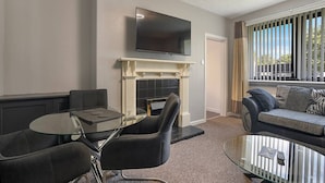 A modern living room with a fireplace, a flat-screen TV mounted above it, a round glass table with chairs, and a sofa with cushions. - Hilton Road Apartment (Aberdeen)