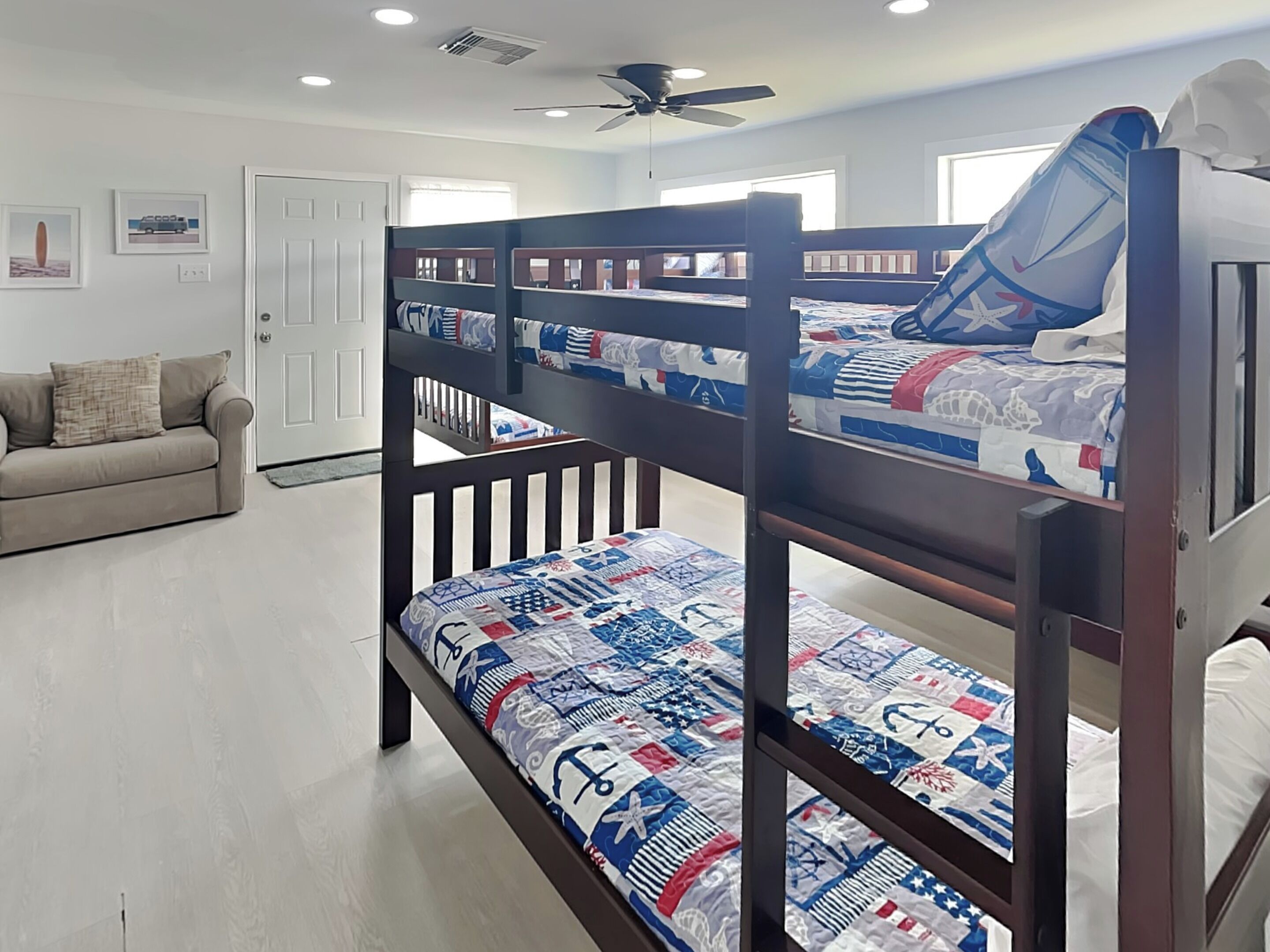 A bunk bed with a quilted bedspread, a ceiling fan, and a door in a room with a light-colored floor and a sofa.