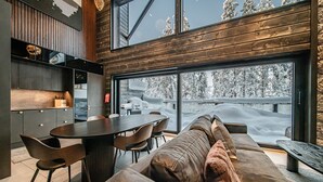 A modern interior with a wooden ceiling, a dining area with a round table and chairs, and a large window overlooking a snowy landscape.