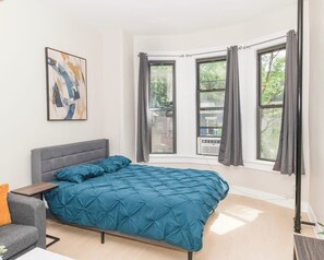 Ein Schlafzimmer mit einem Bett, einem Sessel, einem Fenster mit Vorhängen und einem Gemälde an der Wand.