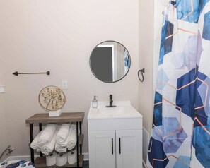 Ein Badezimmer mit einem weißen Waschbecken, einem runden Spiegel, einem Handtuchhalter mit gefalteten Handtüchern und einem Duschvorhang mit geometrischem Muster.