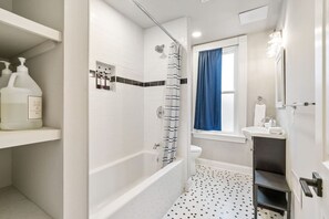 A bathroom with a bathtub, toilet, and sink. There are shelves with toiletries and a window with a curtain. - 4 Story Home|Triple King|Backyard|UPMC Shadyside (Pittsburgh)