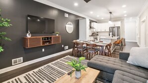 Ein modernes Wohnzimmer mit dunkler Wand, einem Wand-Fernseher, einer Holz-Unterhaltungseinheit, einem Essbereich mit Tisch und Stühlen sowie einer Küche im Hintergrund.