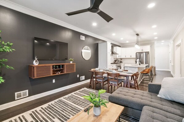 A modern living room with a dark wall, a mounted TV, a wooden entertainment unit, a dining area with a table and chairs, and a kitchen in the background. - 4 Story Home|Triple King|Backyard|UPMC Shadyside (Pittsburgh)