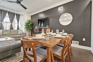 A modern dining area with a wooden table set for four, surrounded by cushioned chairs. A flat-screen TV is mounted on the dark wall above the table. - 4 Story Home|Triple King|Backyard|UPMC Shadyside (Pittsburgh)