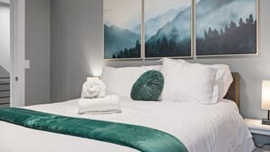 Una cama bien hecha con sábanas blancas y un plaid verde, una mesita de noche con una lámpara, y una pared con tres cuadros de paisajes.