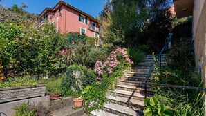 A residential building with a staircase leading to a balcony, surrounded by lush greenery and flowering plants.