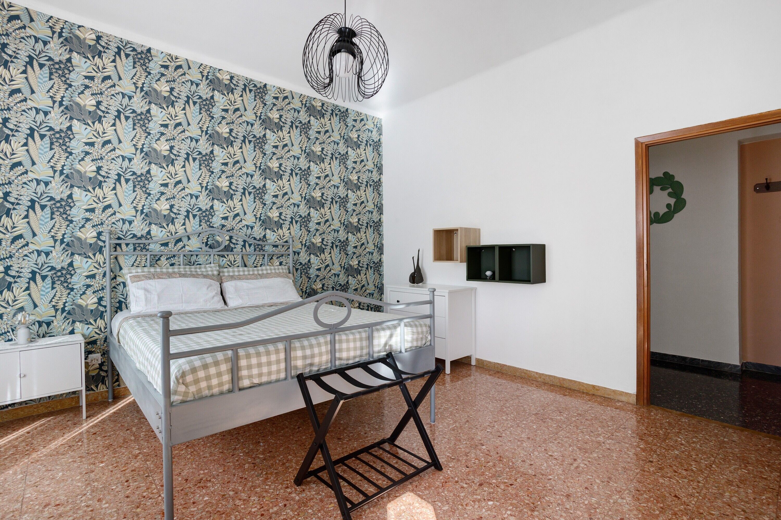 Une chambre à coucher avec un cadre de lit en métal, une table de chevet blanche, un téléviseur fixé au mur et un papier peint à motifs.