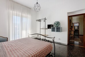 A bedroom with a bed, a hanging light fixture, a coat rack, and a wall-mounted shelf.