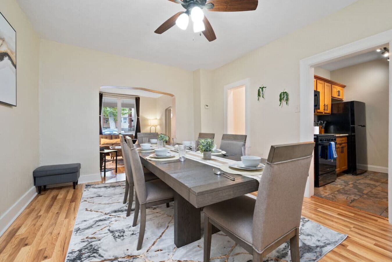 A dining area with a long table, chairs, and a ceiling fan. There is a kitchen visible in the background with wooden cabinets and a black refrigerator.