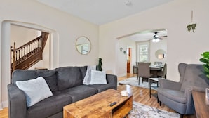 A modern living room with a grey sofa, a wooden coffee table, and a grey armchair. There is a staircase to the left and a dining area in the background. - 3 Story home w/ 11 beds! | Close to Pitt & CMU! (Pittsburgh)