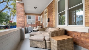 A covered patio area with wicker furniture, a brick wall, and a window with blinds. - 3 Story home w/ 11 beds! | Close to Pitt & CMU! (Pittsburgh)