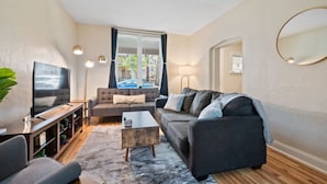 A modern living room with a sofa set, a coffee table, a TV, and a large window with curtains. - 3 Story home w/ 11 beds! | Close to Pitt & CMU! (Pittsburgh)