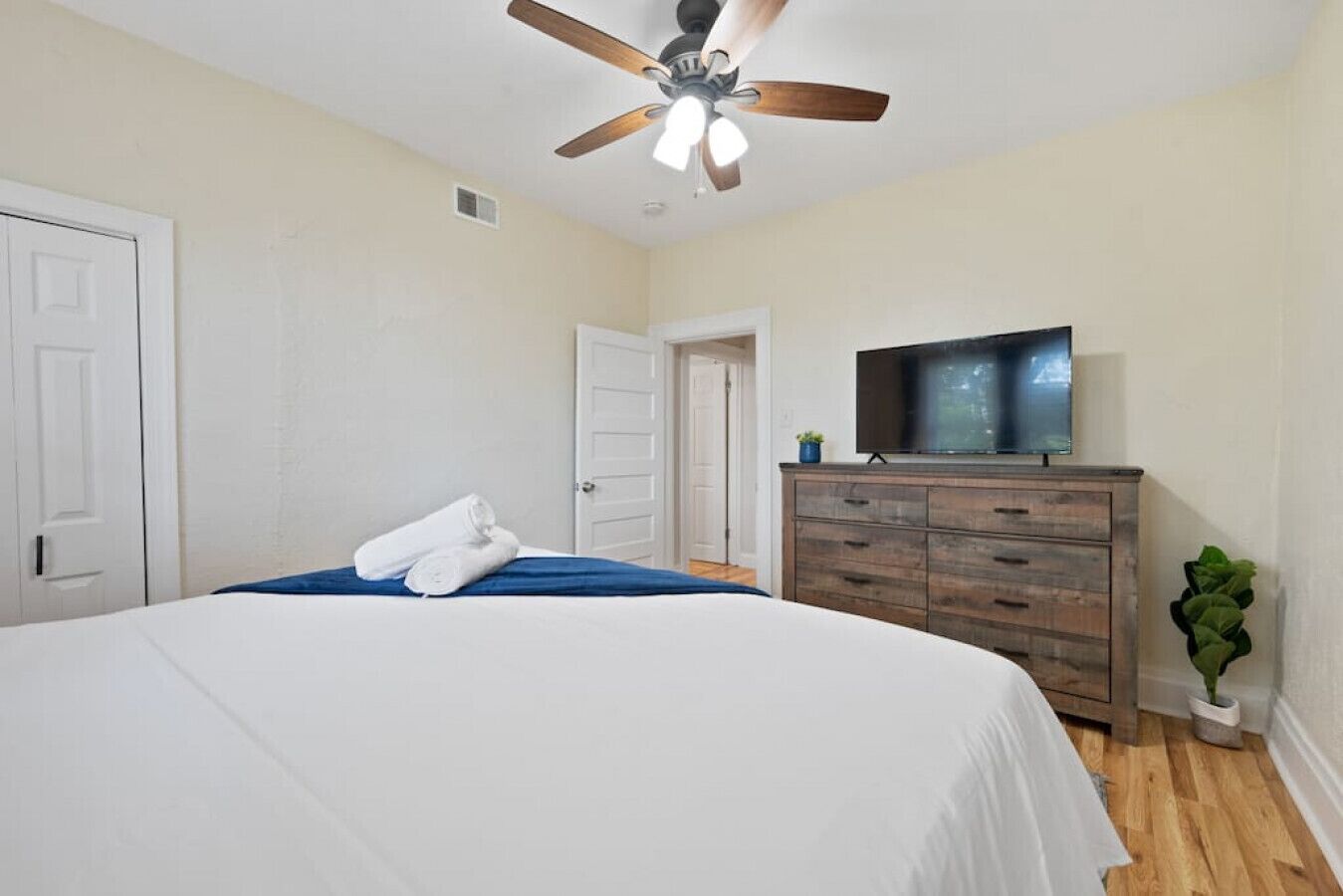 A bedroom with a large bed, a ceiling fan, a TV on a wooden dresser, and a door leading to another room.