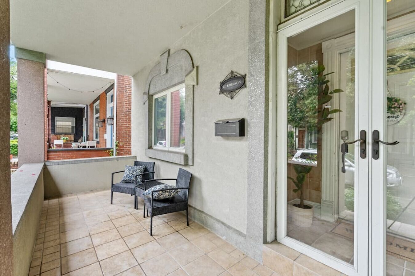 A covered outdoor seating area with two chairs and a small table, a mailbox, and a door with glass panels.