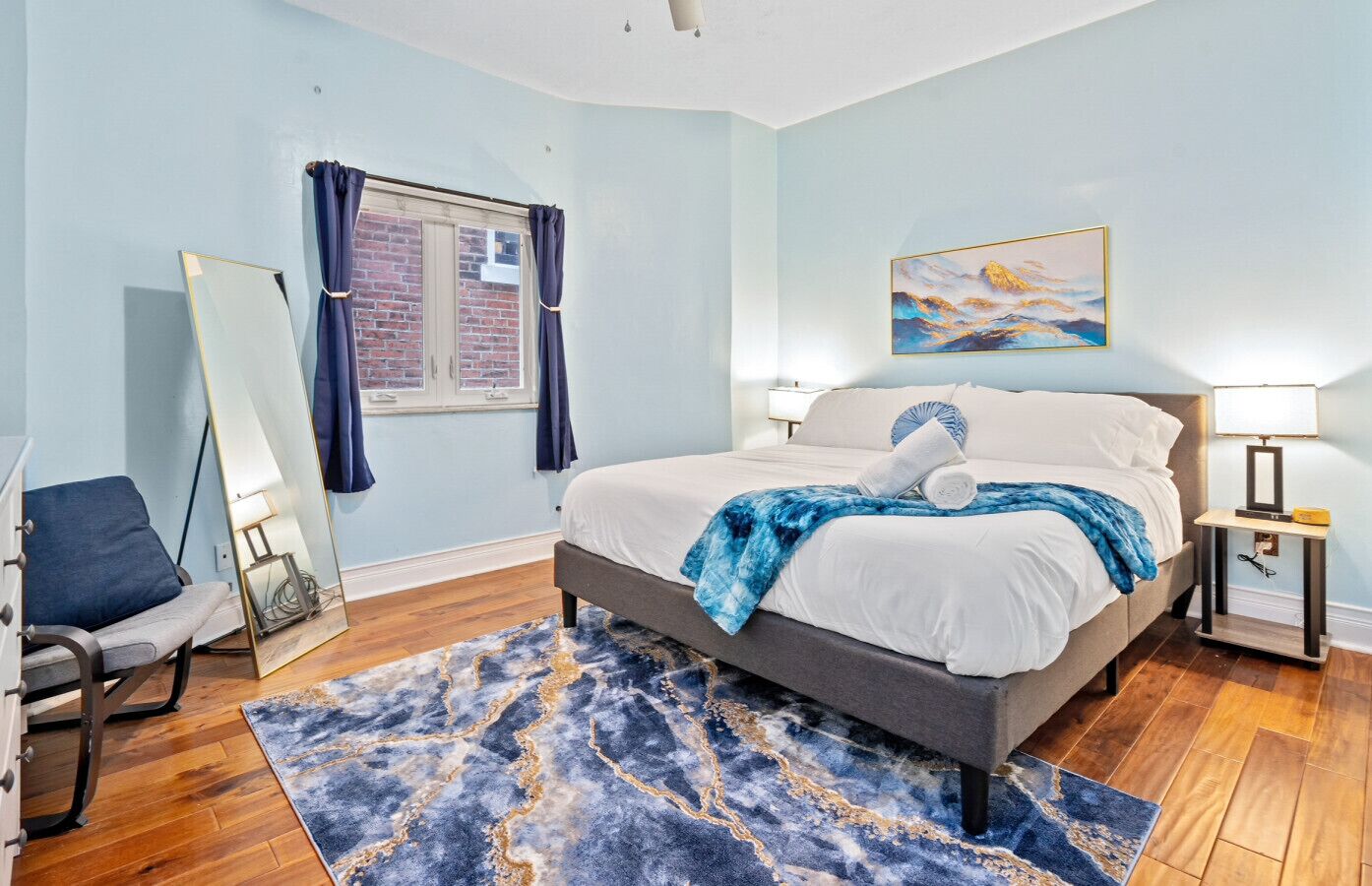 A bedroom with a bed, a chair, a mirror, a painting, and a lamp.