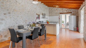 Une cuisine aux murs de pierre et au plafond en bois, un coin repas avec une table dressée pour quatre personnes, et une vue sur l’extérieur grâce à une porte vitrée.