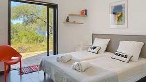 Une chambre d’hôtel moderne dotée d’une grande porte-fenêtre coulissante donnant sur un balcon, d’un fauteuil rouge, de deux lits avec du linge de lit blanc et d’une petite étagère avec des livres et un objet décoratif.