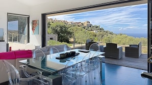 Un espace de repas moderne avec une table et des chaises en verre, un canapé et une vue sur une ville située sur une colline.
