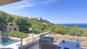 Une terrasse en bois avec une piscine, des meubles en osier et une vue sur la mer et les bâtiments situés sur la colline.