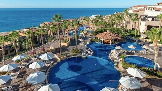 A resort with a large central pool, palm trees, and a view of the ocean.