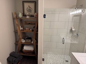 Un baño con ducha, un estante de madera con toallas, un cuadro enmarcado y un letrero que dice 'hogar'.