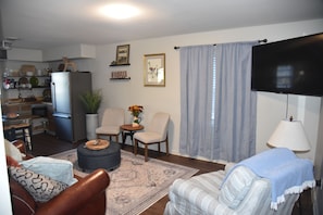 Ein Wohnzimmer mit Sofa, Sessel und Hocker, ein Essbereich mit Tisch und Stühlen, eine Küche mit Geräten und ein an der Wand befestigtes Fernsehgerät.