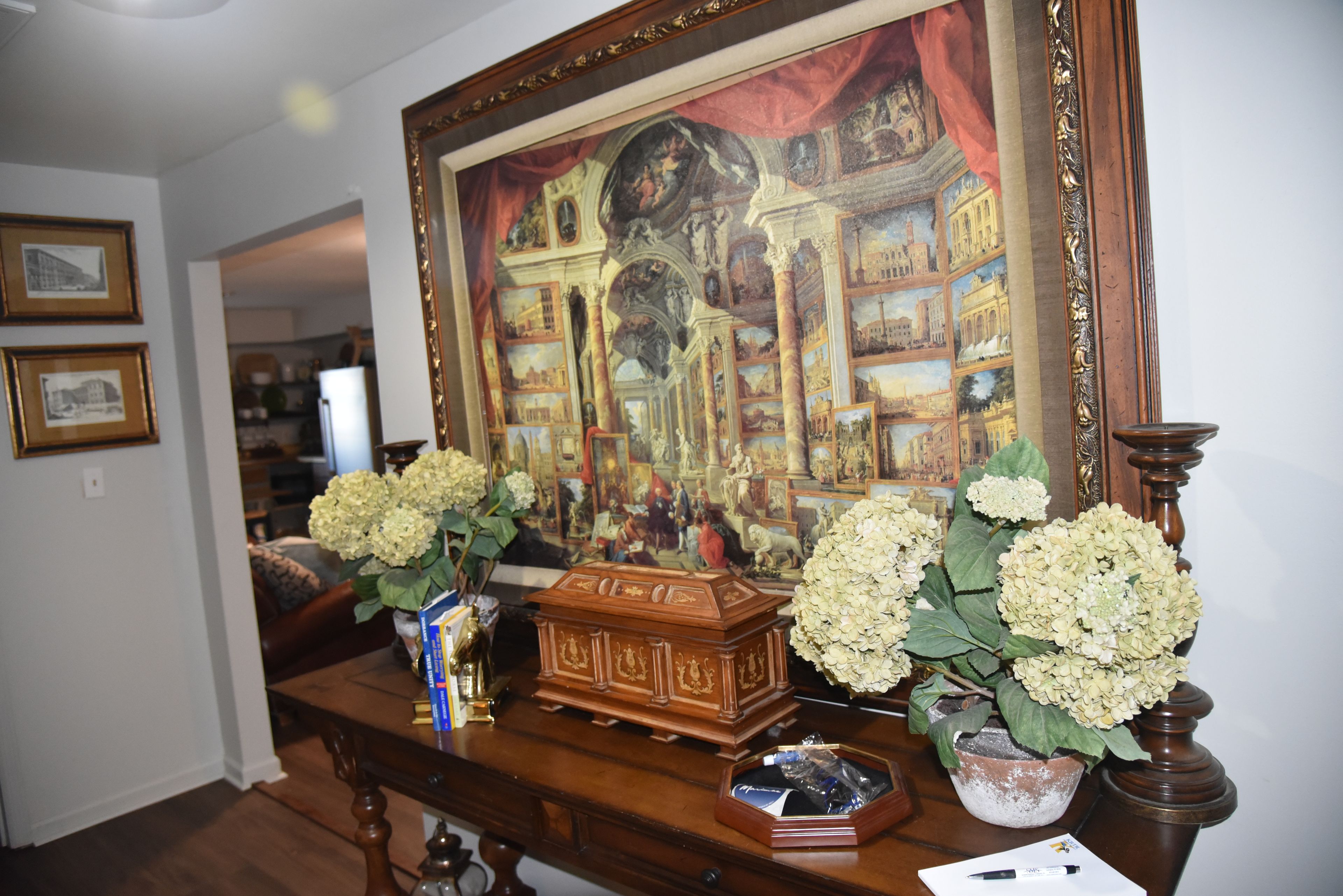A room with a large framed painting, a wooden chest, and two vases with white flowers on a wooden table.