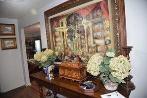 Una habitación con un cuadro grande enmarco, un cofre de madera y dos jarrones con flores blancas sobre una mesa de madera.