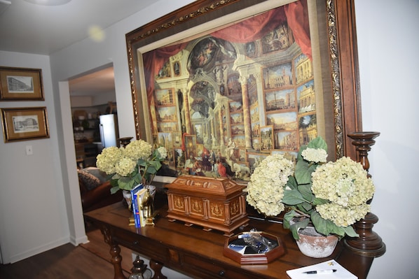 A room with a large framed painting, a wooden chest, and two vases with white flowers on a wooden table.