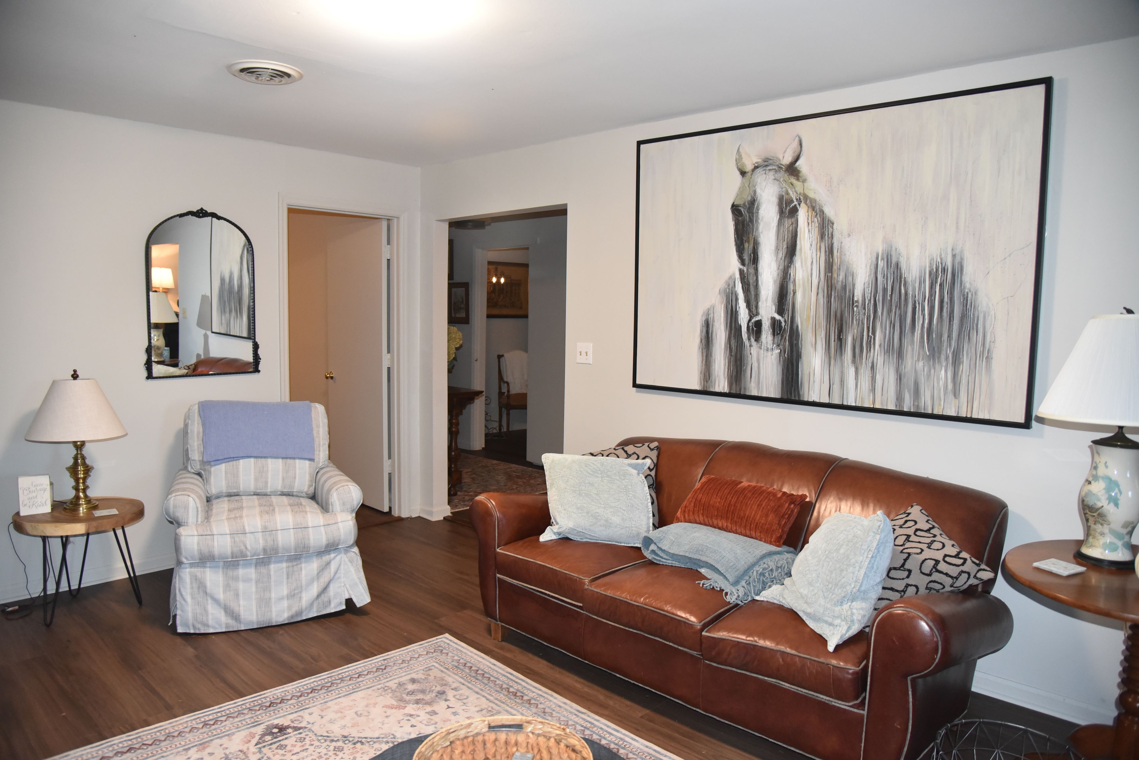 Una sala de estar con un sofá de cuero marrón, un sillón a cuadros, un tapete, una pintura de caballos y un espejo.