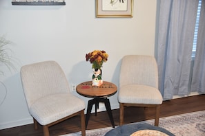 Zwei Polstersessel mit einem kleinen runden Tisch dazwischen, eine Vase mit Blumen und ein gerahmtes Pflanzenbild an der Wand.