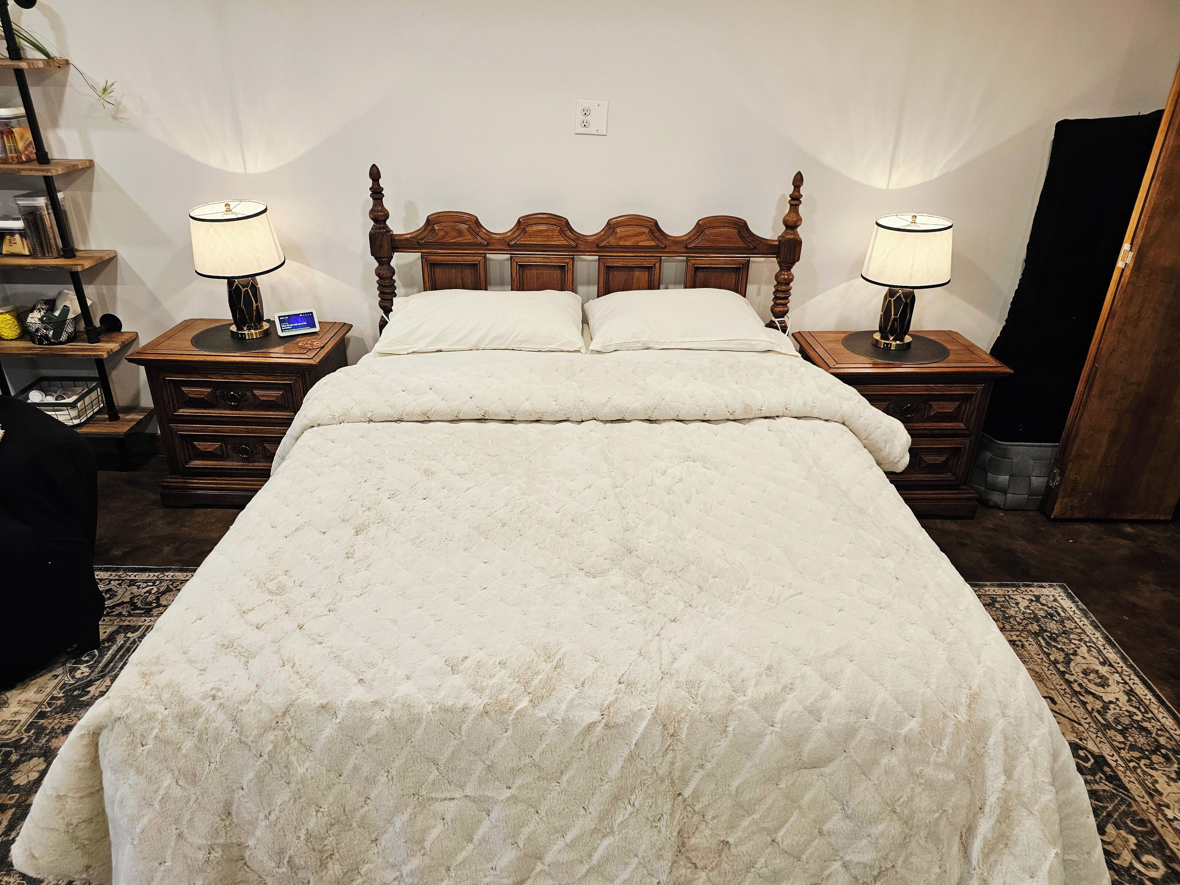 A neatly made bed with a quilted white duvet, two bedside lamps, a wooden nightstand with drawers, and a small table with a lamp and a digital device.