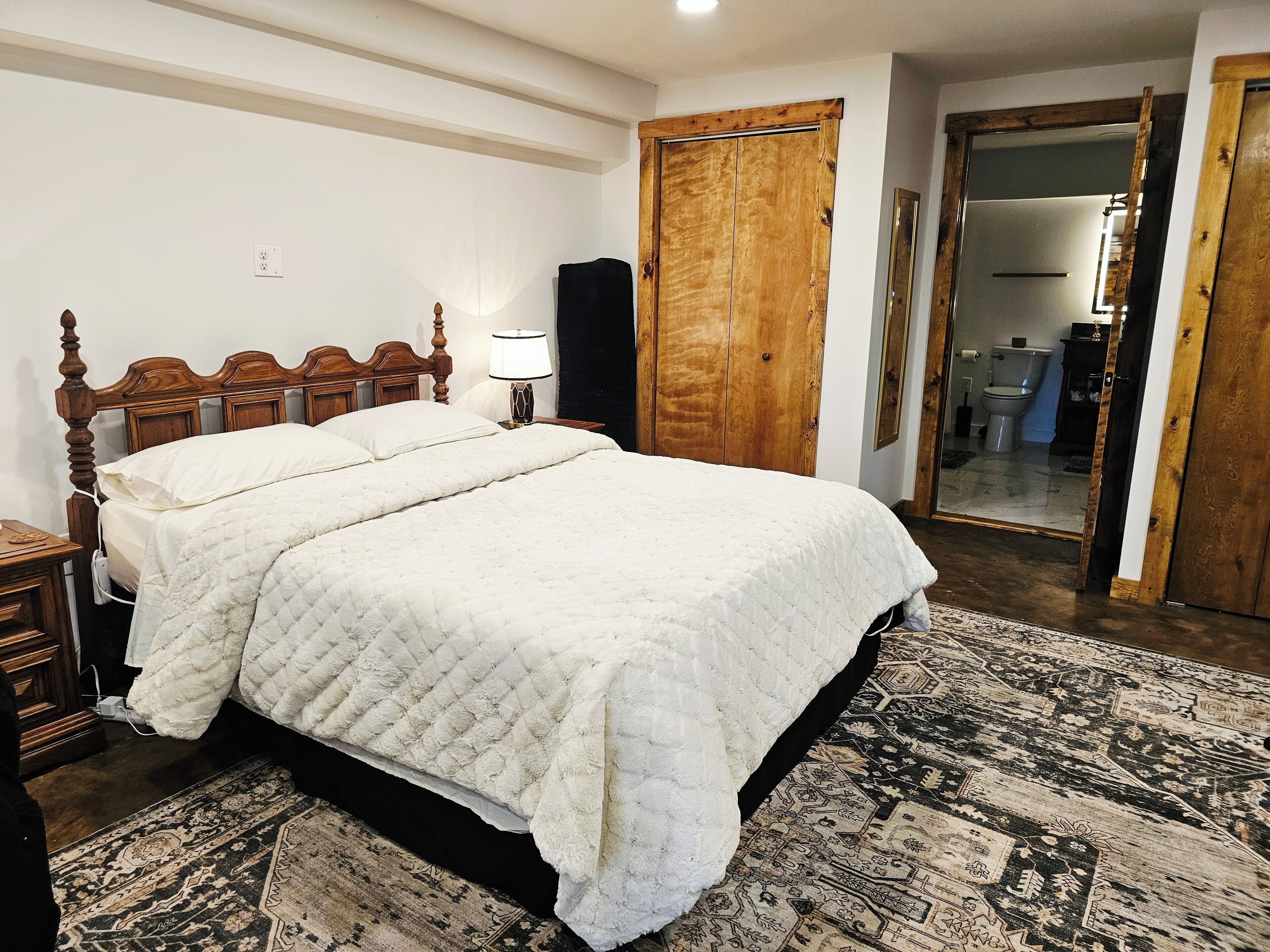 Un dormitorio con una cama de madera, una mesita de noche con lámpara, un espejo grande y una alfombra estampada.