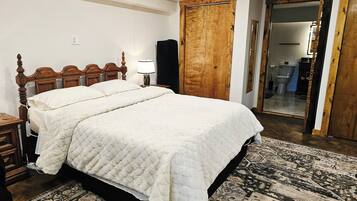 Un dormitorio con una cama de madera, una mesita de noche con lámpara, un espejo grande y una alfombra estampada.