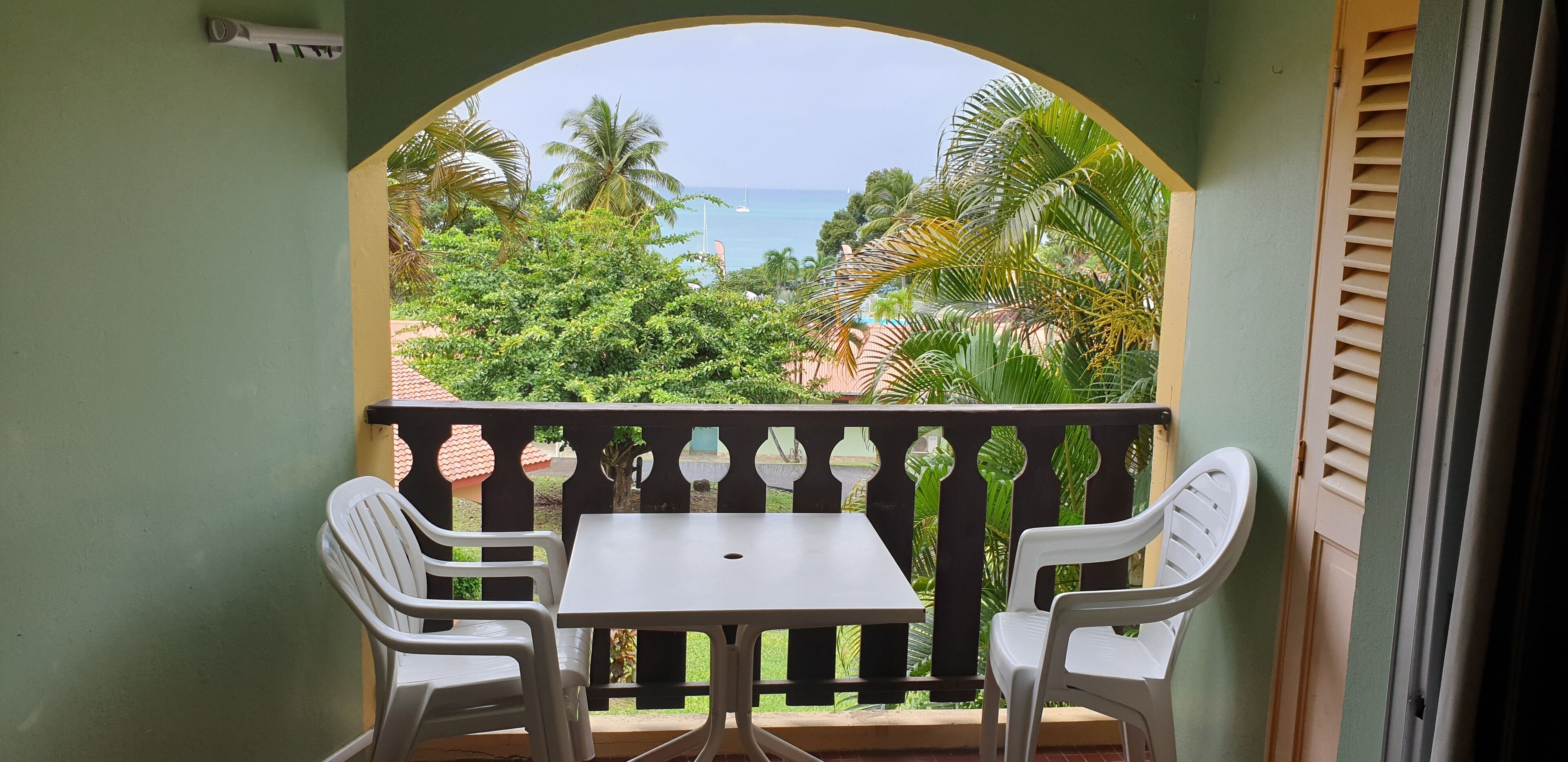 A balcony with two white plastic chairs and a table, overlooking a beach with palm trees and a clear sky.