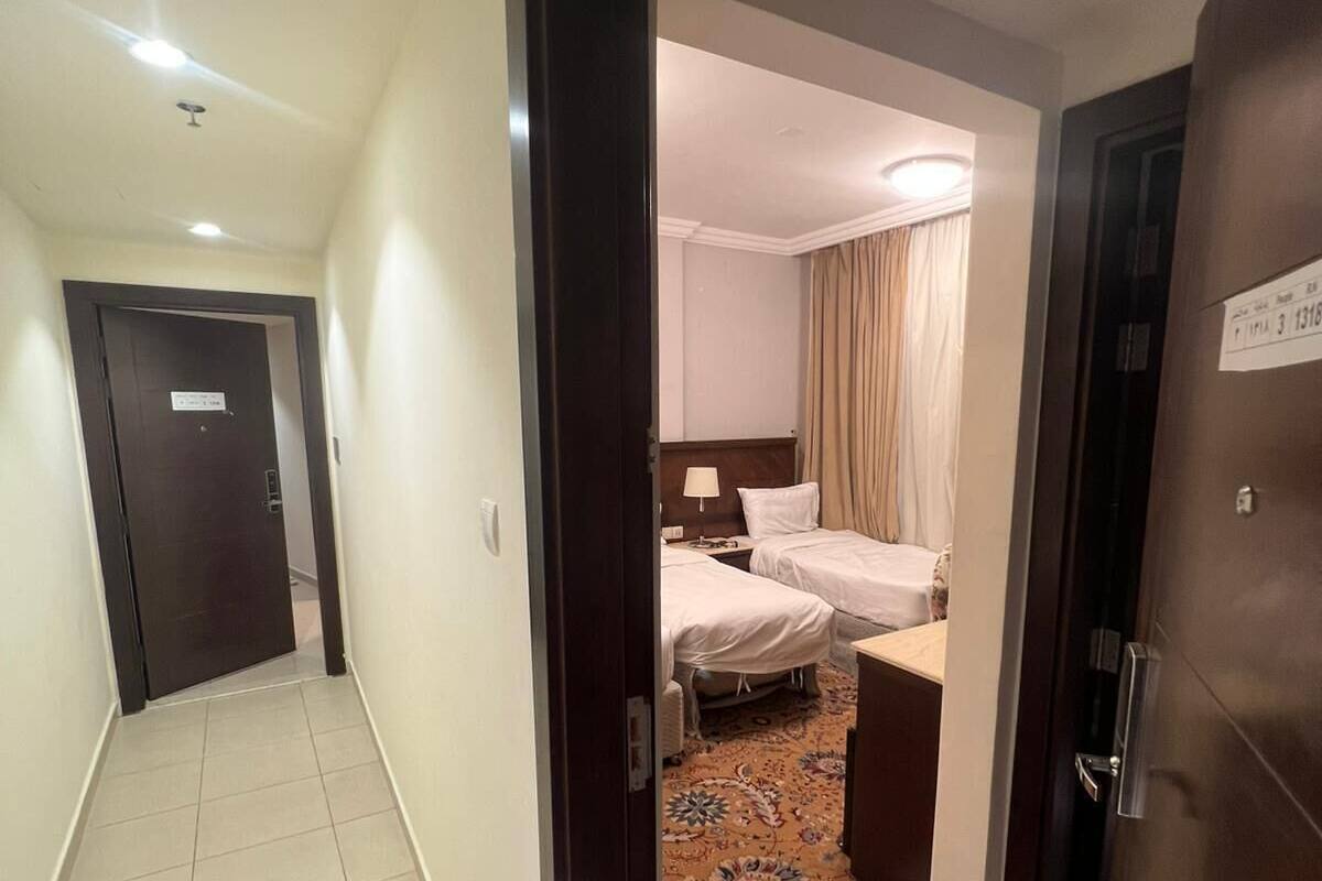 Ein Hotelkorridor mit einem Teppichzimmer, das zwei Betten, einen Nachttisch und ein Fenster mit Vorhängen zeigt.